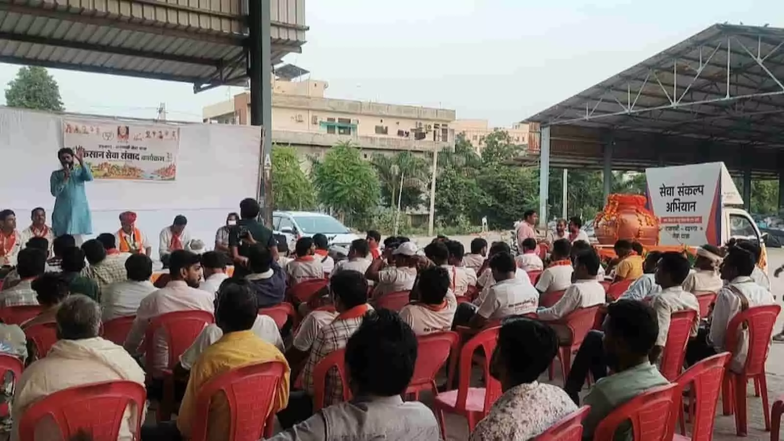 तस्वीर में एक शेड के नीचे बने मंच पर एक व्यक्ति भाषण दे रहा है और सामने कुर्सियों पर लोग बैठे हैं। तस्वीर में एक शेड के नीचे बने मंच पर एक व्यक्ति भाषण दे रहा है और सामने कुर्सियों पर लोग बैठे हैं।