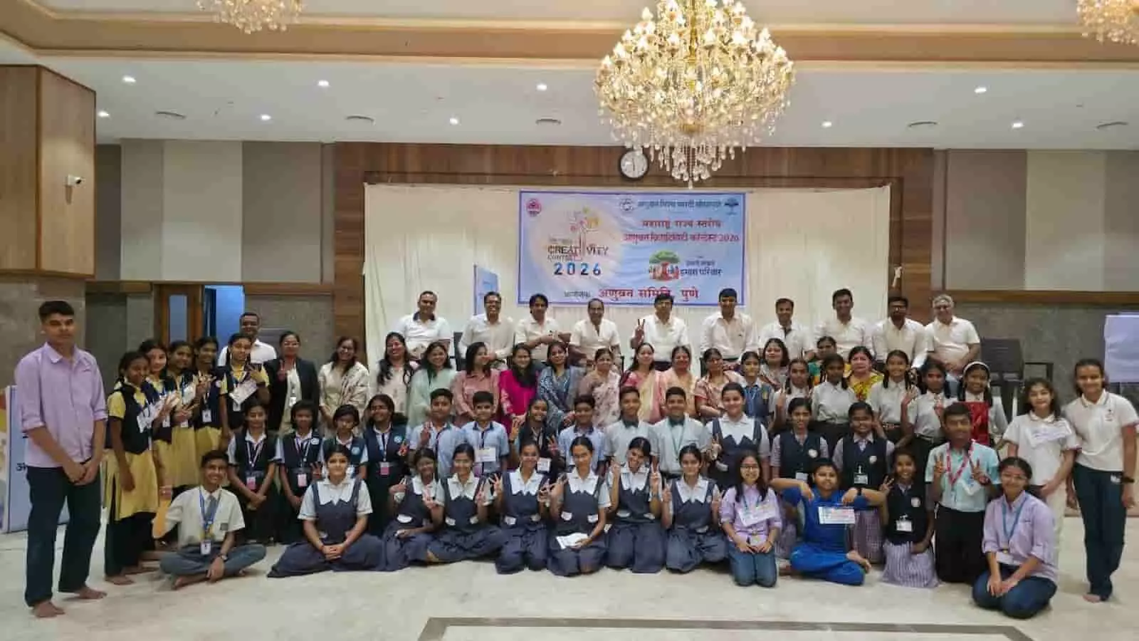 तस्वीर में दिख रही एक बड़े हॉल में मंच के सामने खड़े बच्चे और आयोजक समूह फोटो दे रहे हैं। तस्वीर में दिख रही एक बड़े हॉल में मंच के सामने खड़े बच्चे और आयोजक समूह फोटो दे रहे हैं।