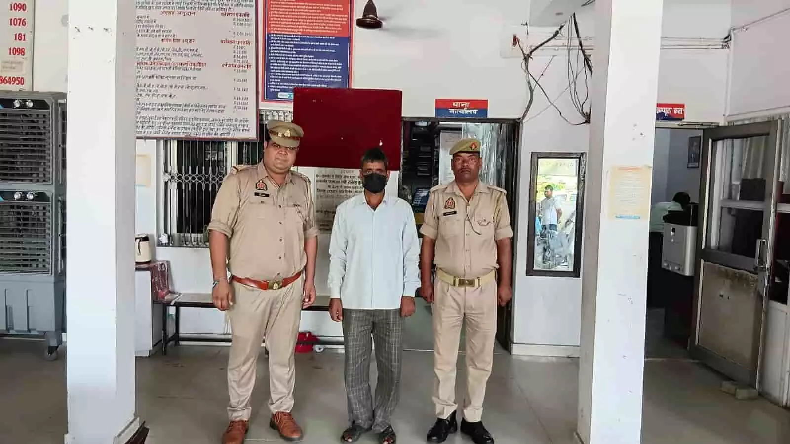 तस्वीर में दो पुलिसकर्मियों के बीच मास्क पहने एक व्यक्ति पुलिस थाने के परिसर में खड़ा है। तस्वीर में दो पुलिसकर्मियों के बीच मास्क पहने एक व्यक्ति पुलिस थाने के परिसर में खड़ा है।