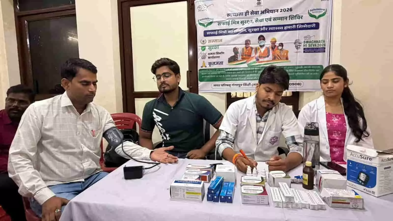 तस्वीर में दिख रही मेज पर दवाइयां रखी हैं और डॉक्टर कर्मचारी का ब्लड प्रेशर जांच रहे हैं। तस्वीर में दिख रही मेज पर दवाइयां रखी हैं और डॉक्टर कर्मचारी का ब्लड प्रेशर जांच रहे हैं।