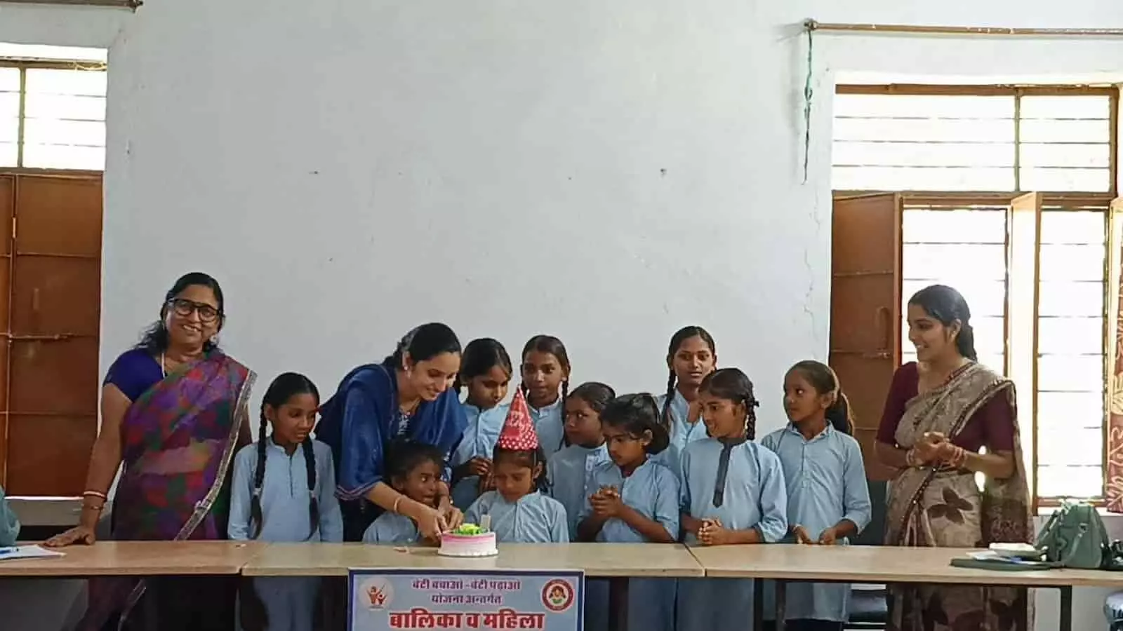 तस्वीर में दिख रही महिलाएं और स्कूल ड्रेस पहनी बालिकाएं टेबल के पास खड़ी होकर केक काट रही हैं। तस्वीर में दिख रही महिलाएं और स्कूल ड्रेस पहनी बालिकाएं टेबल के पास खड़ी होकर केक काट रही हैं।
