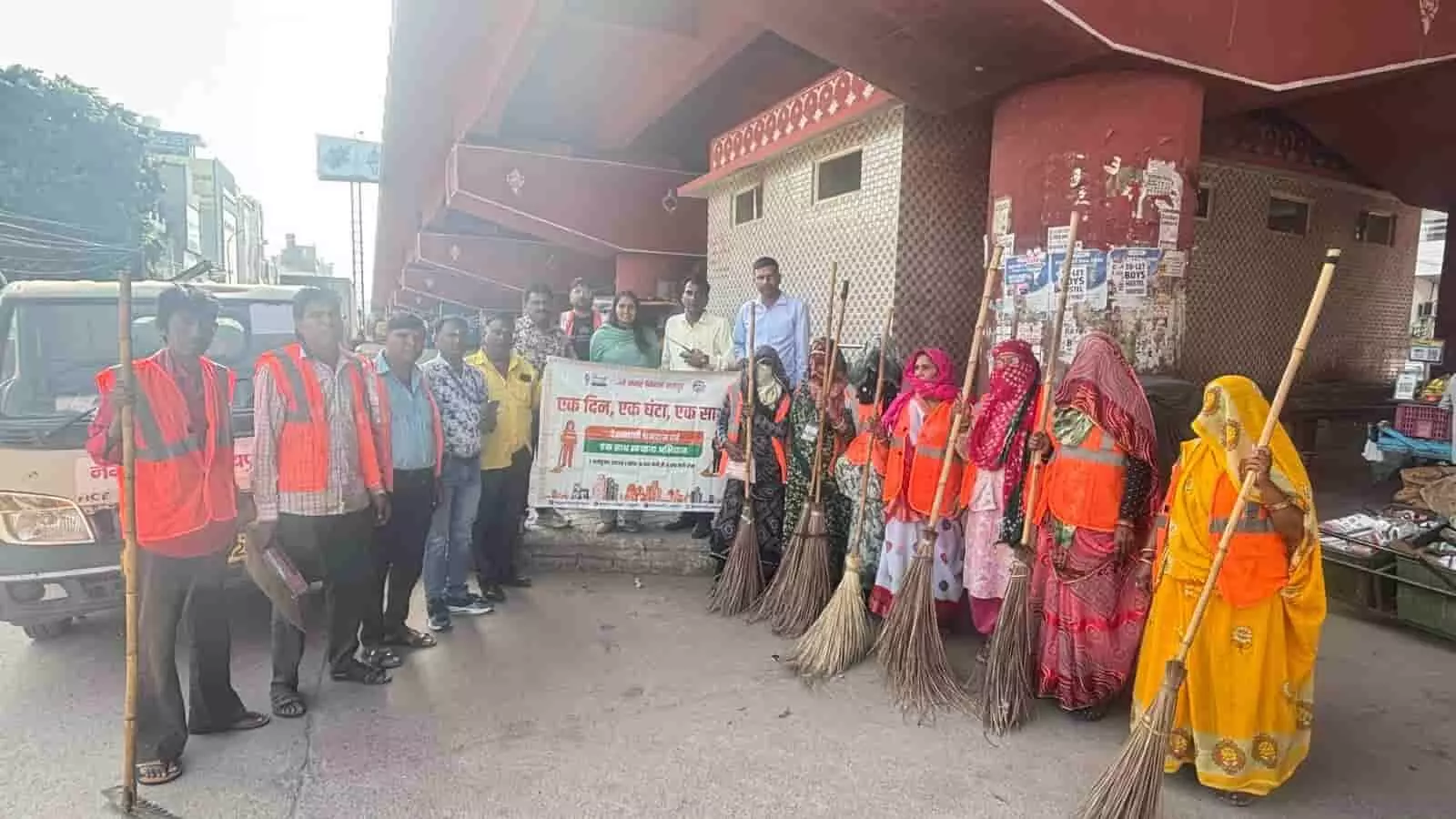 तस्वीर में बाएं से दाएं खड़े अधिकारी, कर्मचारी और झाड़ू थामे महिलाएं बैनर के साथ दिख रही हैं। तस्वीर में बाएं से दाएं खड़े अधिकारी, कर्मचारी और झाड़ू थामे महिलाएं बैनर के साथ दिख रही हैं।