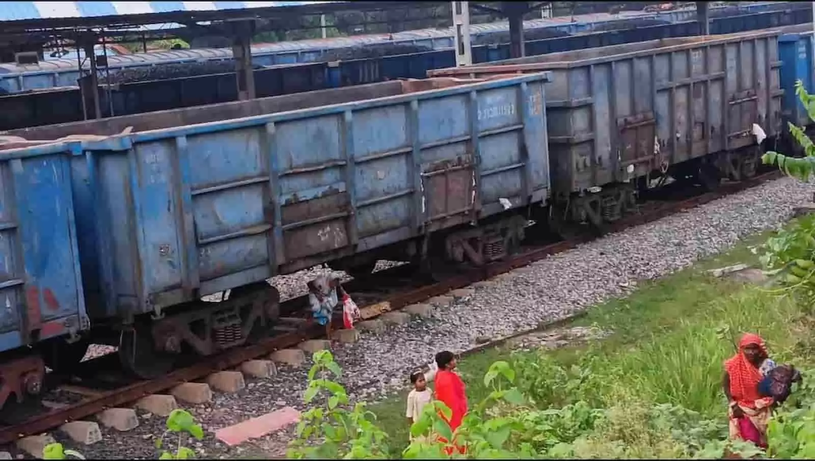 तस्वीर में रेलवे स्टेशन पर नीली मालगाड़ी के नीचे और पास से लोग पटरियां पार करते दिख रहे हैं। तस्वीर में रेलवे स्टेशन पर नीली मालगाड़ी के नीचे और पास से लोग पटरियां पार करते दिख रहे हैं।