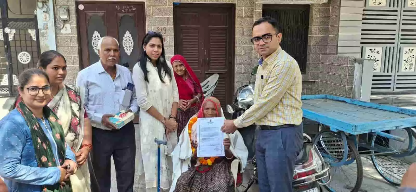 तस्वीर में बाएं से दाएं लोग एक बुजुर्ग महिला के साथ खड़े हैं और अधिकारी उन्हें दस्तावेज दे रहे हैं। तस्वीर में बाएं से दाएं लोग एक बुजुर्ग महिला के साथ खड़े हैं और अधिकारी उन्हें दस्तावेज दे रहे हैं।