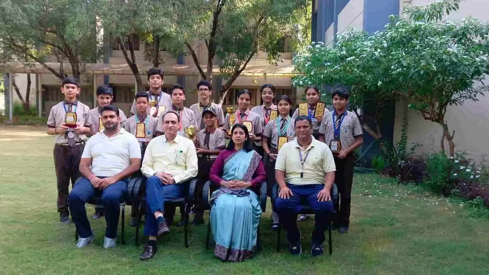 तस्वीर में आगे कुर्सियों पर बैठे शिक्षकों के पीछे हाथ में ट्रॉफियां लिए विद्यार्थी खड़े हैं। तस्वीर में आगे कुर्सियों पर बैठे शिक्षकों के पीछे हाथ में ट्रॉफियां लिए विद्यार्थी खड़े हैं।