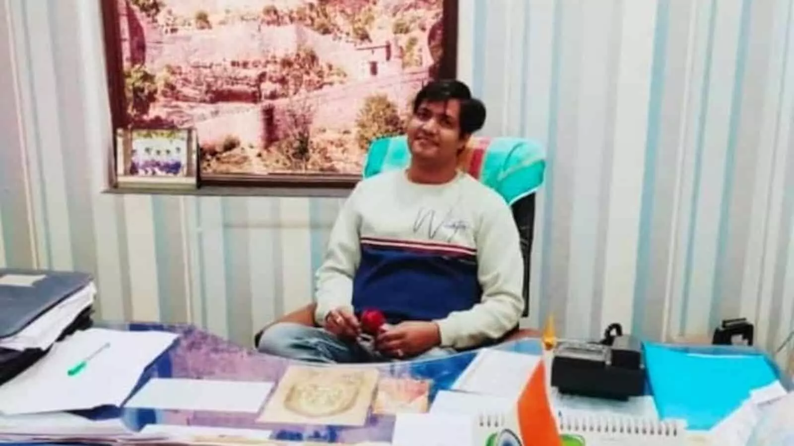 तस्वीर में एक युवक कार्यालय के केबिन में मेज के पीछे कुर्सी पर बैठकर फोटो खिंचवा रहा है। तस्वीर में एक युवक कार्यालय के केबिन में मेज के पीछे कुर्सी पर बैठकर फोटो खिंचवा रहा है।
