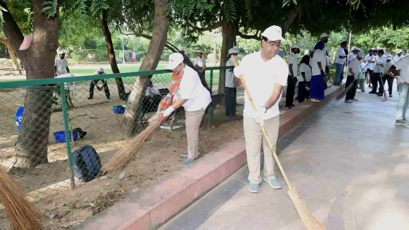 तस्वीर में बाईं ओर एक महिला और दाईं ओर एक व्यक्ति पार्क परिसर में झाड़ू से सफाई कर रहे हैं। तस्वीर में बाईं ओर एक महिला और दाईं ओर एक व्यक्ति पार्क परिसर में झाड़ू से सफाई कर रहे हैं।