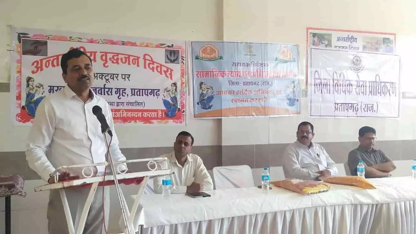 तस्वीर में बाएं से दाएं एक अधिकारी पॉडियम पर बोलते हुए और मेज पर तीन अन्य व्यक्ति बैठे दिख रहे हैं। तस्वीर में बाएं से दाएं एक अधिकारी पॉडियम पर बोलते हुए और मेज पर तीन अन्य व्यक्ति बैठे दिख रहे हैं।