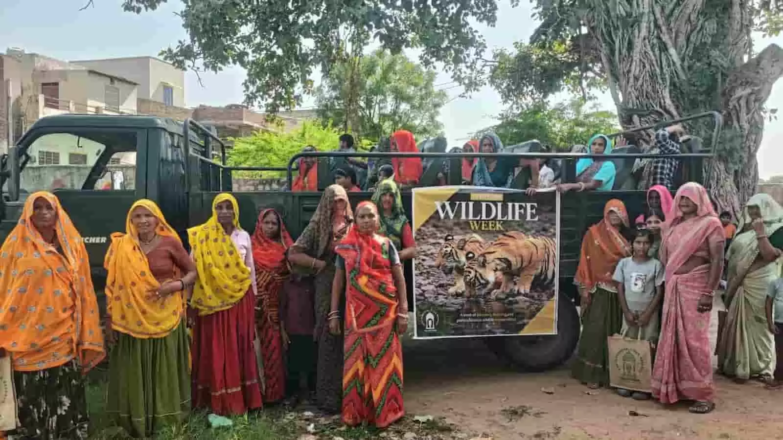 तस्वीर में पारंपरिक पोशाक पहने ग्रामीण महिलाएं खुली सफारी गाड़ी के पास वन्यजीव पोस्टर के साथ खड़ी हैं। तस्वीर में पारंपरिक पोशाक पहने ग्रामीण महिलाएं खुली सफारी गाड़ी के पास वन्यजीव पोस्टर के साथ खड़ी हैं।