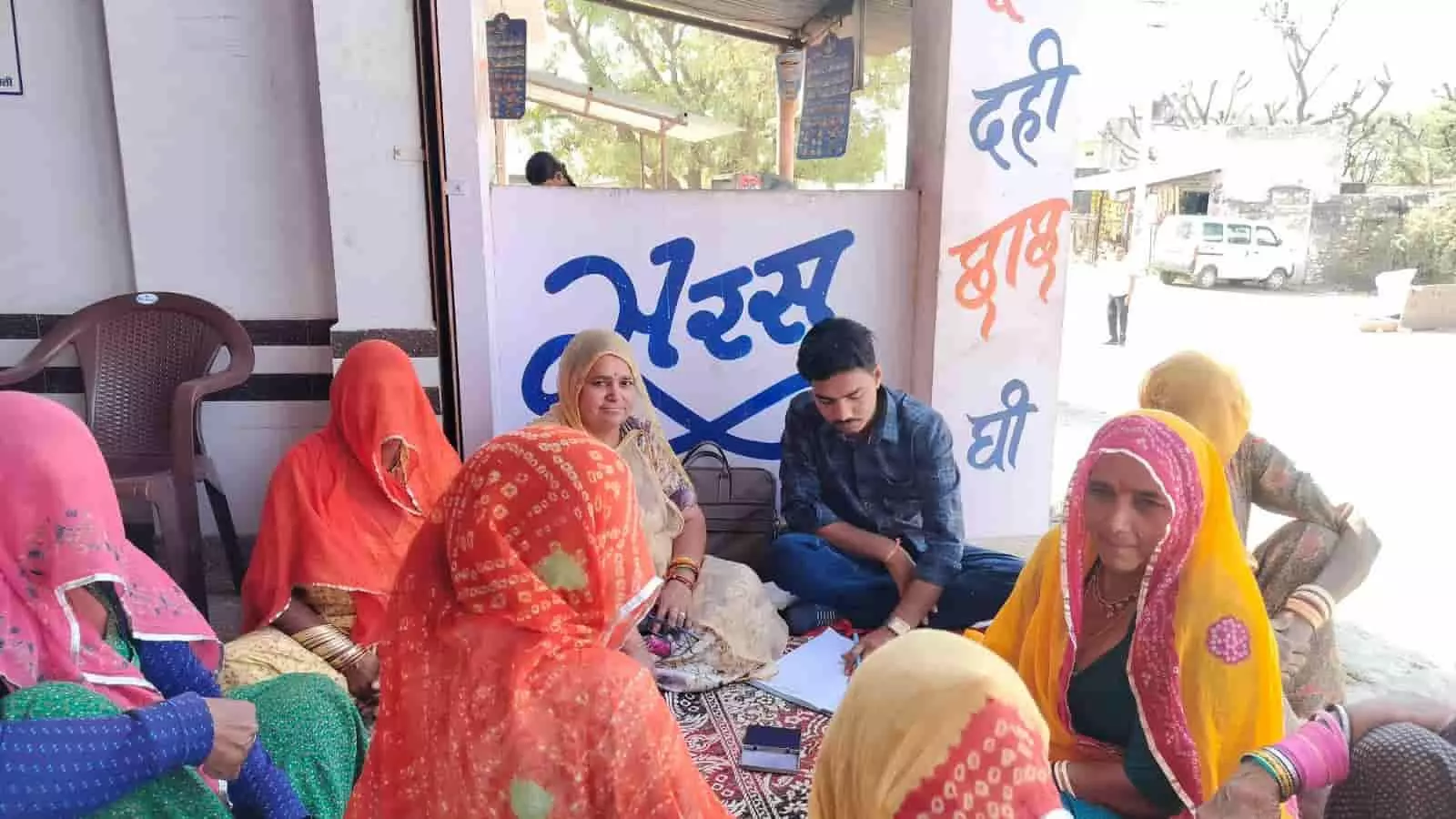 सरस दुग्ध केंद्र के सामने जमीन पर बैठकर बातचीत करती ग्रामीण महिलाएं और एक व्यक्ति। सरस दुग्ध केंद्र के सामने जमीन पर बैठकर बातचीत करती ग्रामीण महिलाएं और एक व्यक्ति।