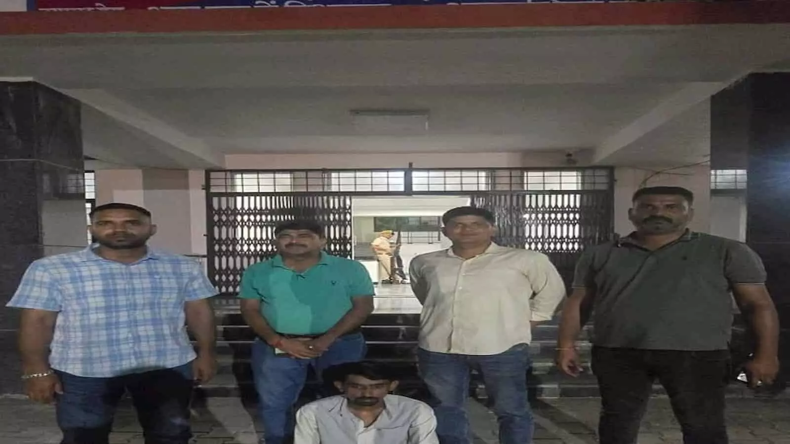 तस्वीर में पुलिस स्टेशन के बाहर खड़े चार पुलिस अधिकारियों के बीच में नीचे आरोपी बैठा दिख रहा है। तस्वीर में पुलिस स्टेशन के बाहर खड़े चार पुलिस अधिकारियों के बीच में नीचे आरोपी बैठा दिख रहा है।