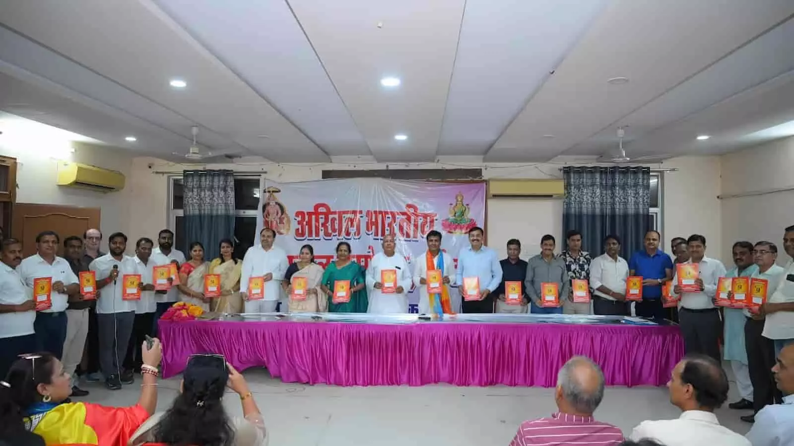 तस्वीर में दिख रही एक हॉल की मंच पर मौजूद कई लोग हाथ में पुस्तिकाएं थामे हुए खड़े हैं। तस्वीर में दिख रही एक हॉल की मंच पर मौजूद कई लोग हाथ में पुस्तिकाएं थामे हुए खड़े हैं।