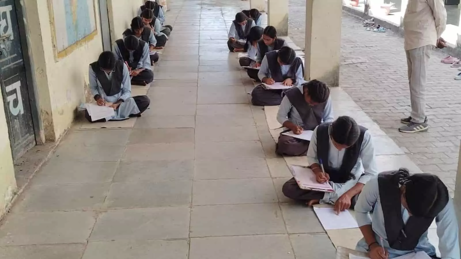 तस्वीर में छात्राएं स्कूल के बरामदे में फर्श पर बैठकर परीक्षा की कॉपियों में उत्तर लिख रही हैं। तस्वीर में छात्राएं स्कूल के बरामदे में फर्श पर बैठकर परीक्षा की कॉपियों में उत्तर लिख रही हैं।