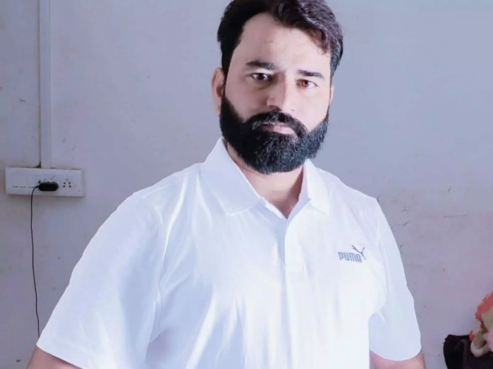 तस्वीर में सफेद पोलो टी-शर्ट और दाढ़ी वाले व्यक्ति कैमरे की ओर देख रहे हैं। तस्वीर में सफेद पोलो टी-शर्ट और दाढ़ी वाले व्यक्ति कैमरे की ओर देख रहे हैं।