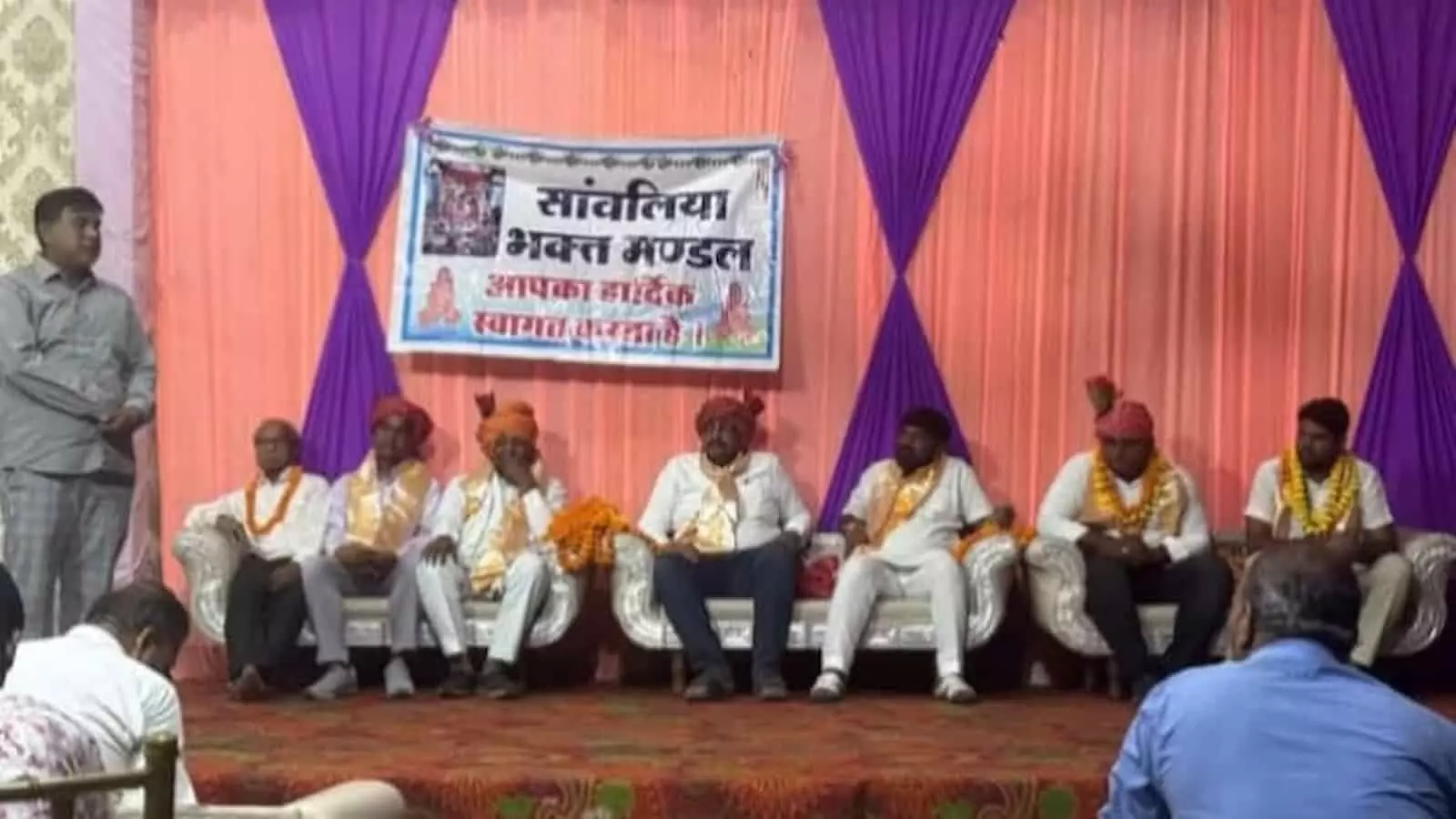 तस्वीर में सांवलिया भक्त मंडल के मंच पर साफा और माला पहने लोग सोफे पर बैठे हुए हैं। तस्वीर में सांवलिया भक्त मंडल के मंच पर साफा और माला पहने लोग सोफे पर बैठे हुए हैं।