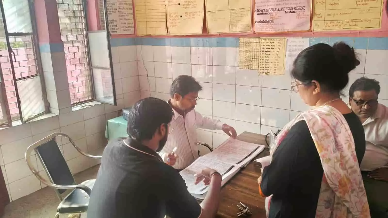 तस्वीर में स्वास्थ्य केंद्र के कमरे में मेज पर खुले रजिस्टर को देखते स्वास्थ्य अधिकारी। तस्वीर में स्वास्थ्य केंद्र के कमरे में मेज पर खुले रजिस्टर को देखते स्वास्थ्य अधिकारी।