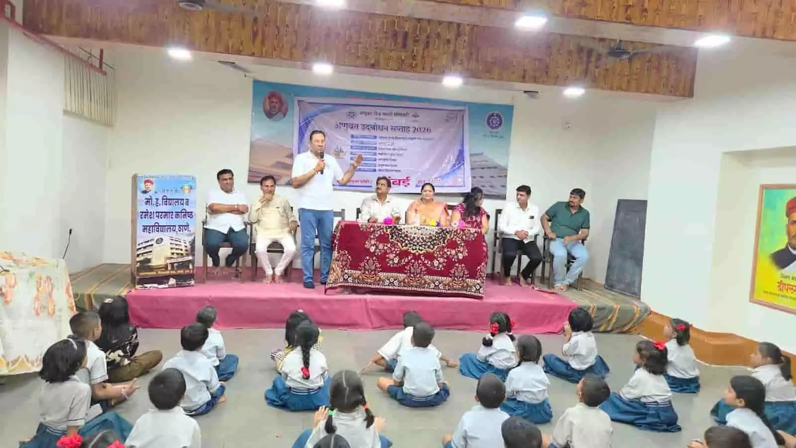 तस्वीर में एक व्यक्ति मंच पर खड़े होकर फर्श पर बैठे स्कूली बच्चों को संबोधित कर रहा है। तस्वीर में एक व्यक्ति मंच पर खड़े होकर फर्श पर बैठे स्कूली बच्चों को संबोधित कर रहा है।