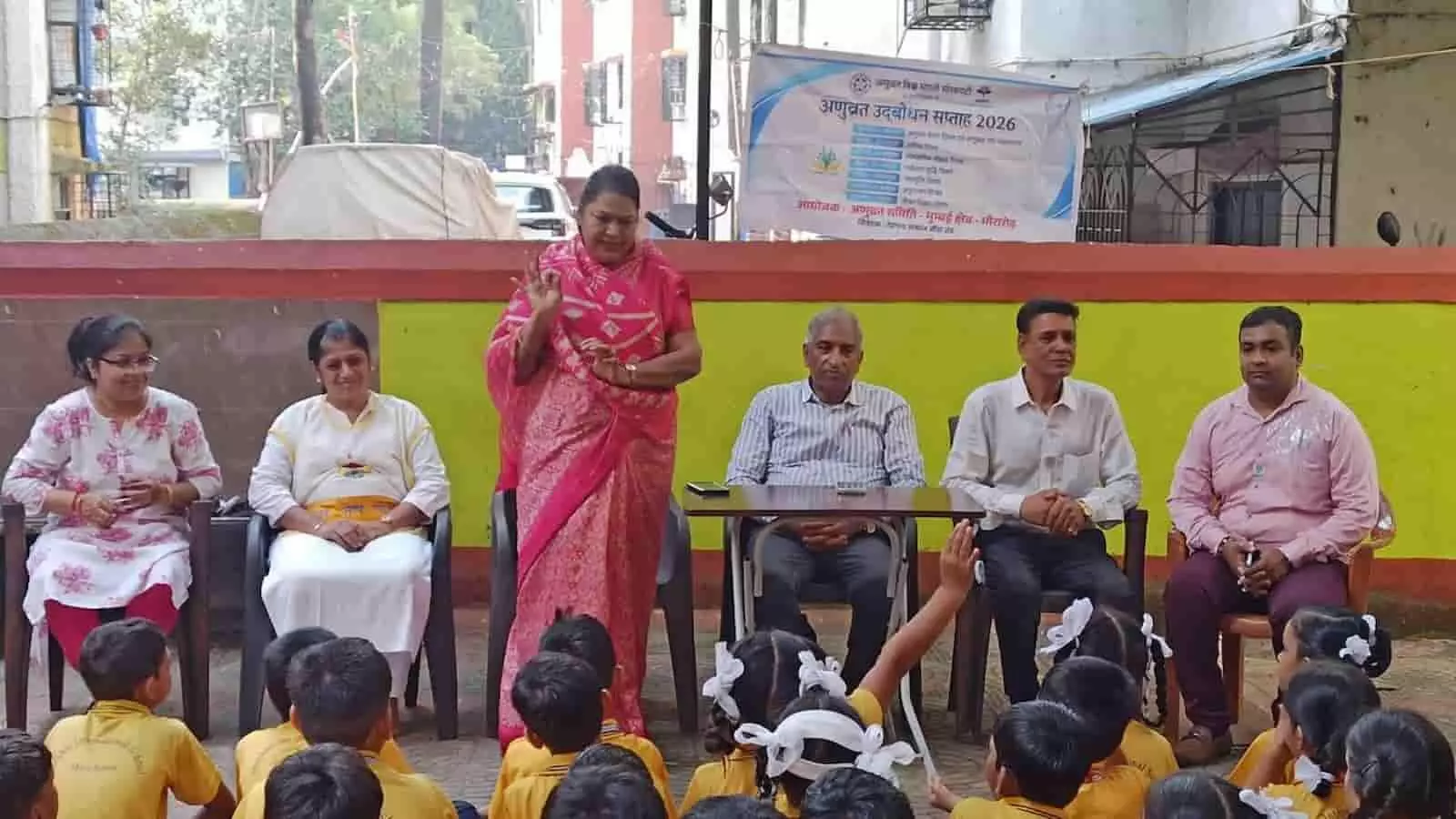 तस्वीर में बाईं से दाईं ओर बैठे अतिथियों के बीच एक महिला बच्चों को मुद्रा समझा रही हैं। तस्वीर में बाईं से दाईं ओर बैठे अतिथियों के बीच एक महिला बच्चों को मुद्रा समझा रही हैं।