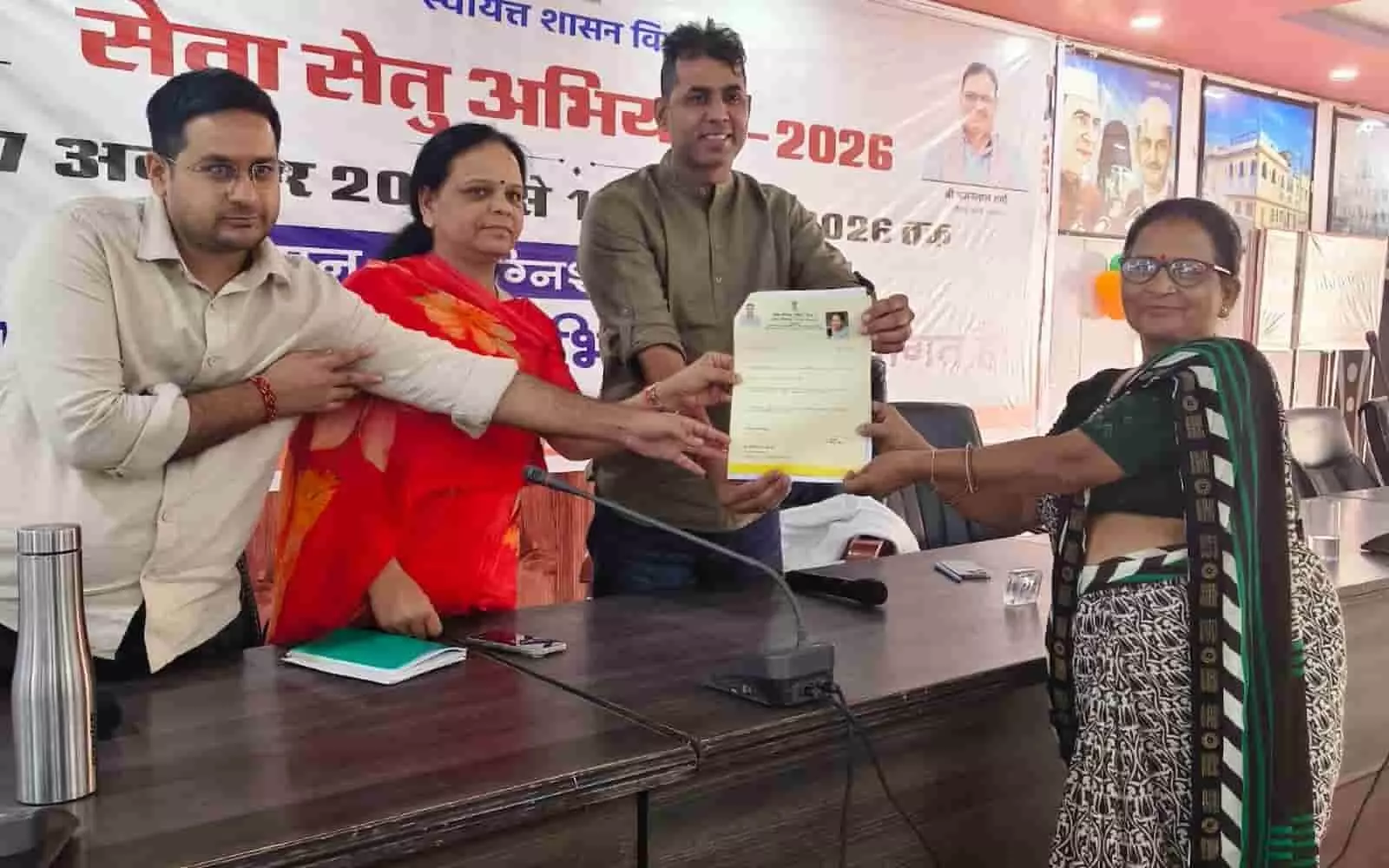 तस्वीर में बाईं ओर खड़े अधिकारी एक महिला को आधिकारिक सरकारी दस्तावेज थमाते दिख रहे हैं। तस्वीर में बाईं ओर खड़े अधिकारी एक महिला को आधिकारिक सरकारी दस्तावेज थमाते दिख रहे हैं।