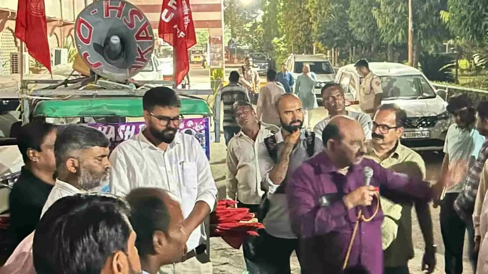 तस्वीर में लाल शर्ट पहने व्यक्ति हाथ में माइक थामे अन्य लोगों के बीच बात रखता दिख रहा है। तस्वीर में लाल शर्ट पहने व्यक्ति हाथ में माइक थामे अन्य लोगों के बीच बात रखता दिख रहा है।