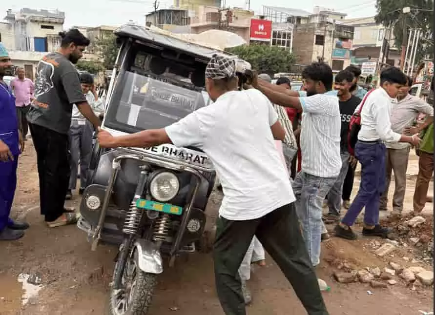 तस्वीर में खराब सड़क पर पलटी हुई ई-रिक्शा को धक्का देकर उठाते कई लोग दिखाई दे रहे हैं। तस्वीर में खराब सड़क पर पलटी हुई ई-रिक्शा को धक्का देकर उठाते कई लोग दिखाई दे रहे हैं।