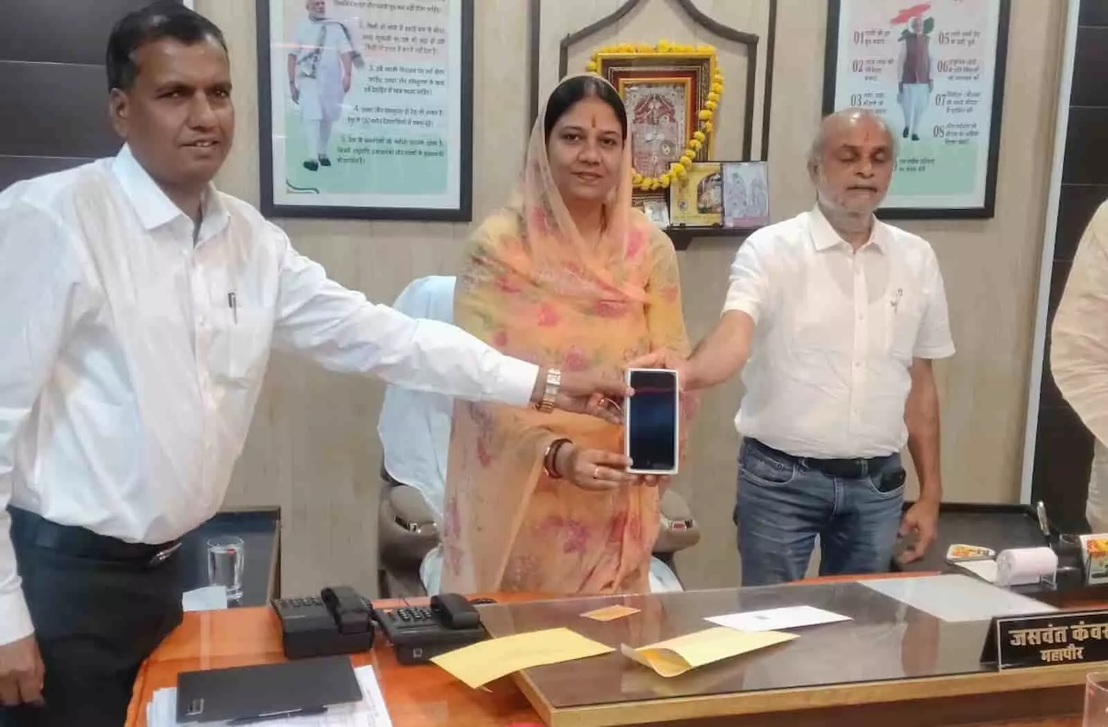तस्वीर में दिख रही महिला और दो पुरुष अधिकारी एक कमरे में मेज के पीछे खड़े होकर मोबाइल दिखाते हैं। तस्वीर में दिख रही महिला और दो पुरुष अधिकारी एक कमरे में मेज के पीछे खड़े होकर मोबाइल दिखाते हैं।