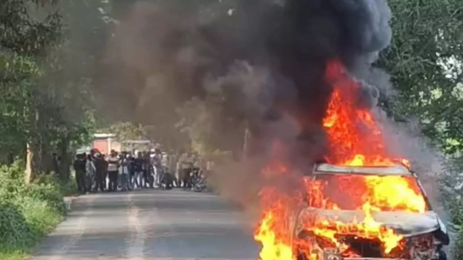 तस्वीर में सड़क पर आग की लपटों से घिरी जलती कार और दूर खड़े लोग दिख रहे हैं। तस्वीर में सड़क पर आग की लपटों से घिरी जलती कार और दूर खड़े लोग दिख रहे हैं।