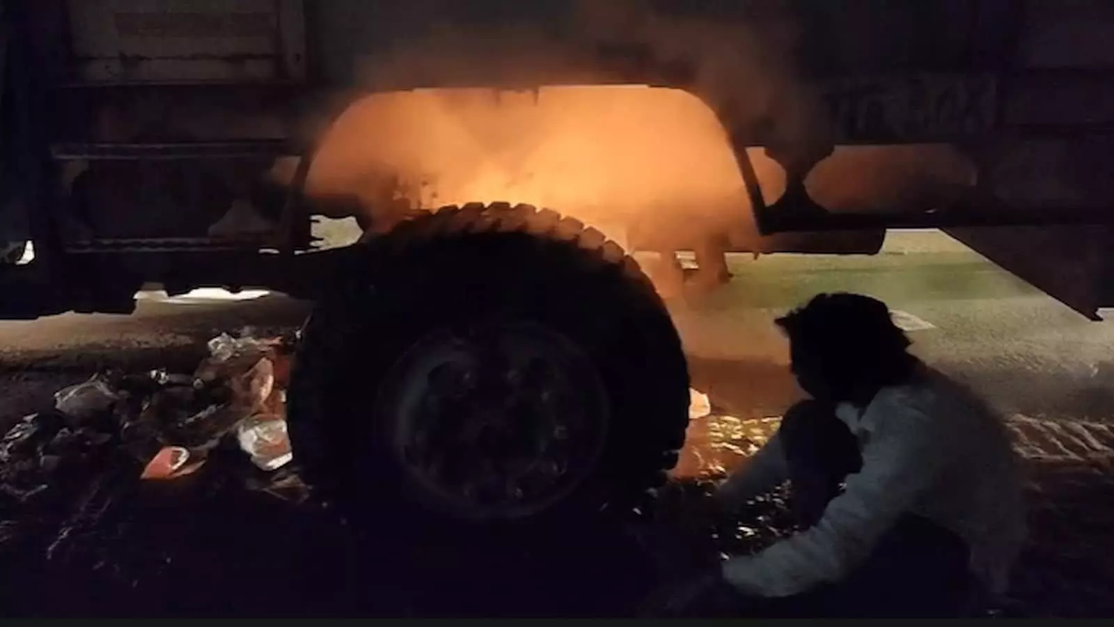 तस्वीर में एटा में लकड़ी से भरे ट्रक के टायर के पास लगी आग और एक व्यक्ति नजर आ रहा है। तस्वीर में एटा में लकड़ी से भरे ट्रक के टायर के पास लगी आग और एक व्यक्ति नजर आ रहा है।