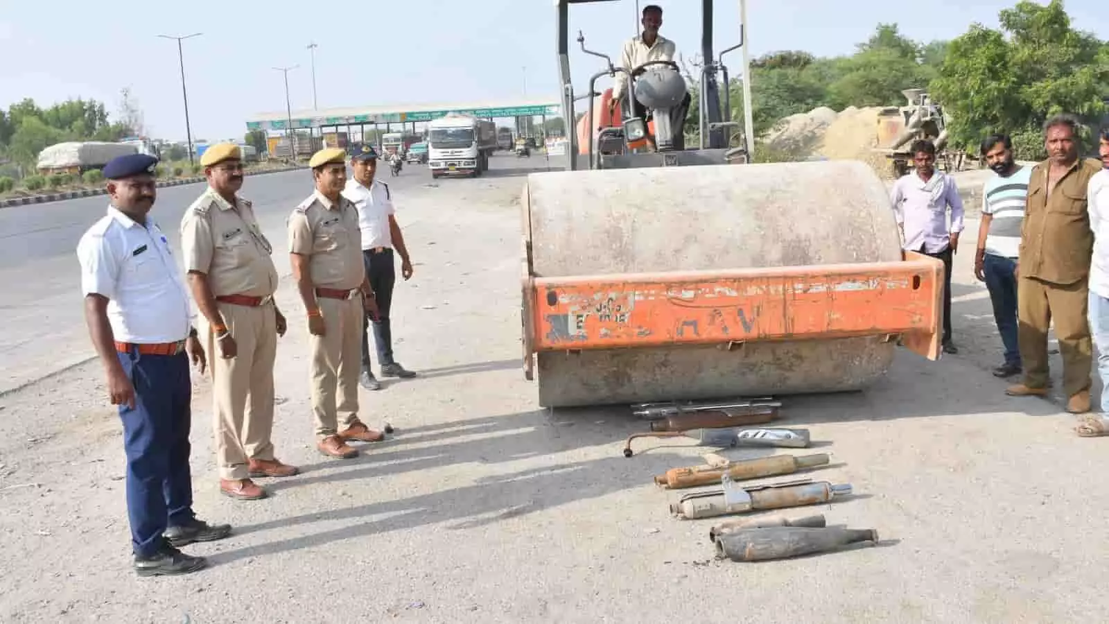 सड़क पर रखे मॉडिफाइड साइलेंसरों के पास पुलिसकर्मी खड़े हैं और रोड रोलर खड़ा है। सड़क पर रखे मॉडिफाइड साइलेंसरों के पास पुलिसकर्मी खड़े हैं और रोड रोलर खड़ा है।