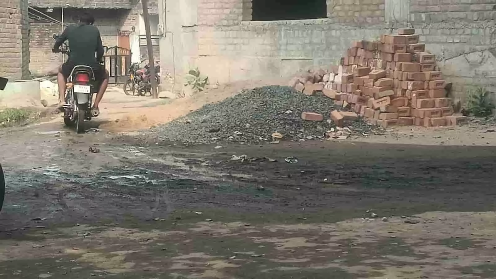 तस्वीर में एक व्यक्ति बाइक से कच्चे रास्ते पर बहते गंदे पानी और पड़े निर्माण सामग्री के पास से गुजर रहा है। तस्वीर में एक व्यक्ति बाइक से कच्चे रास्ते पर बहते गंदे पानी और पड़े निर्माण सामग्री के पास से गुजर रहा है।
