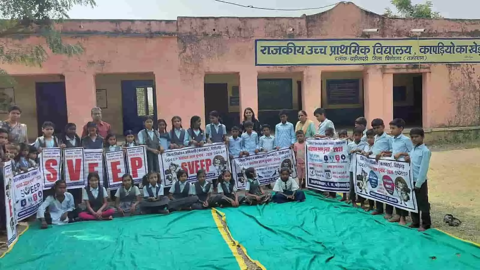 तस्वीर में राजकीय उच्च प्राथमिक विद्यालय कापड़िया का खेड़ा के बाहर स्वीप पोस्टर थामे बच्चे और शिक्षक दिख रहे हैं। तस्वीर में राजकीय उच्च प्राथमिक विद्यालय कापड़िया का खेड़ा के बाहर स्वीप पोस्टर थामे बच्चे और शिक्षक दिख रहे हैं।
