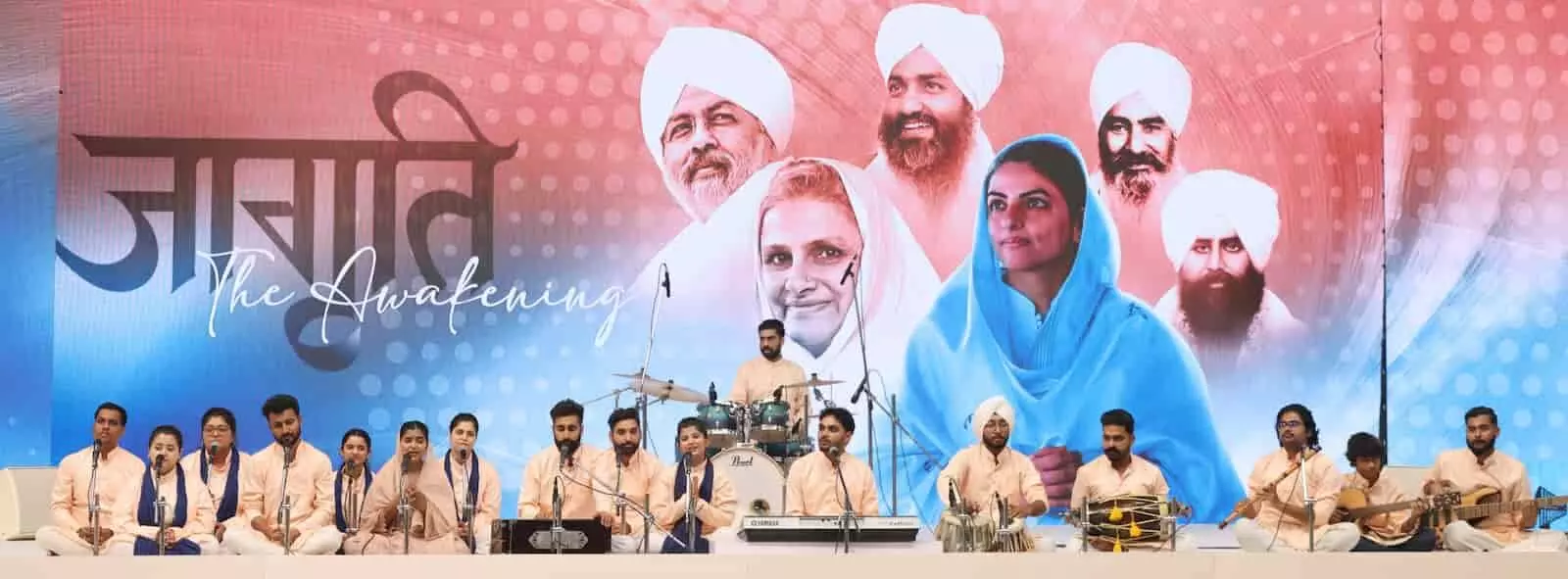 तस्वीर में निरंकारी संत समागम के मंच पर कलाकार और पृष्ठभूमि में गुरुओं के चित्र दिख रहे हैं।