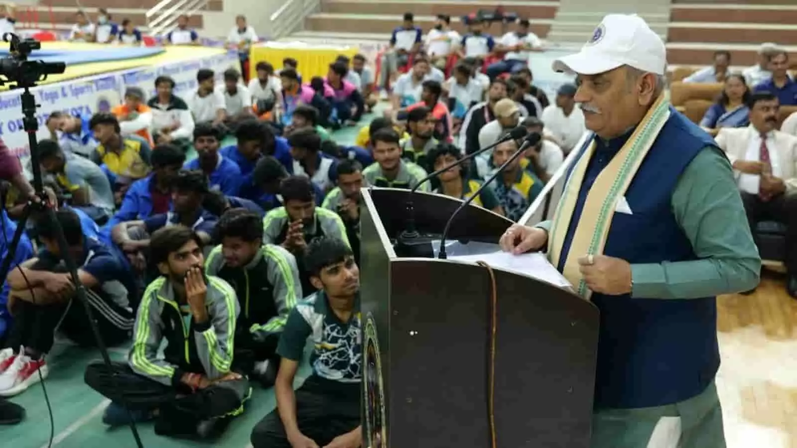 तस्वीर में एक वक्ता मंच के पॉडियम से विश्वविद्यालय के इंडोर हॉल में मौजूद खिलाड़ियों को संबोधित कर रहा है। तस्वीर में एक वक्ता मंच के पॉडियम से विश्वविद्यालय के इंडोर हॉल में मौजूद खिलाड़ियों को संबोधित कर रहा है।