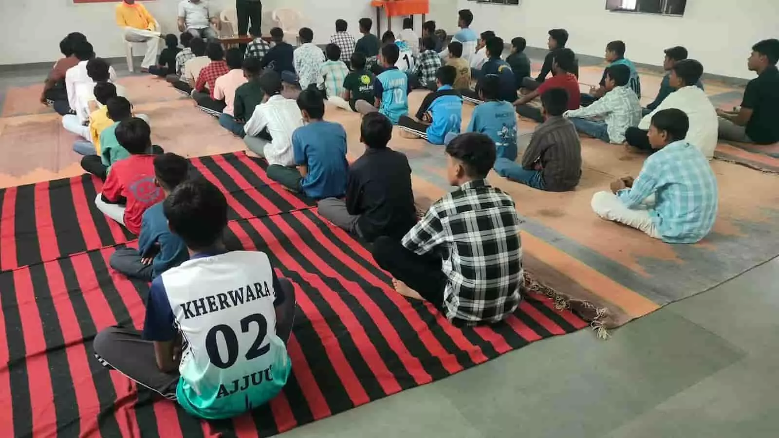 तस्वीर में श्री हरि ओम जनजाति छात्रावास खेरवाड़ा के हॉल में जमीन पर बैठे युवा और अतिथि दिख रहे हैं।